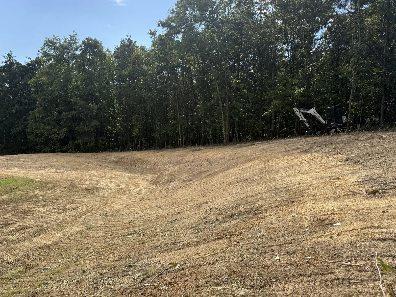 Wide graded bank below a tree line with the mini excavator parked at the top