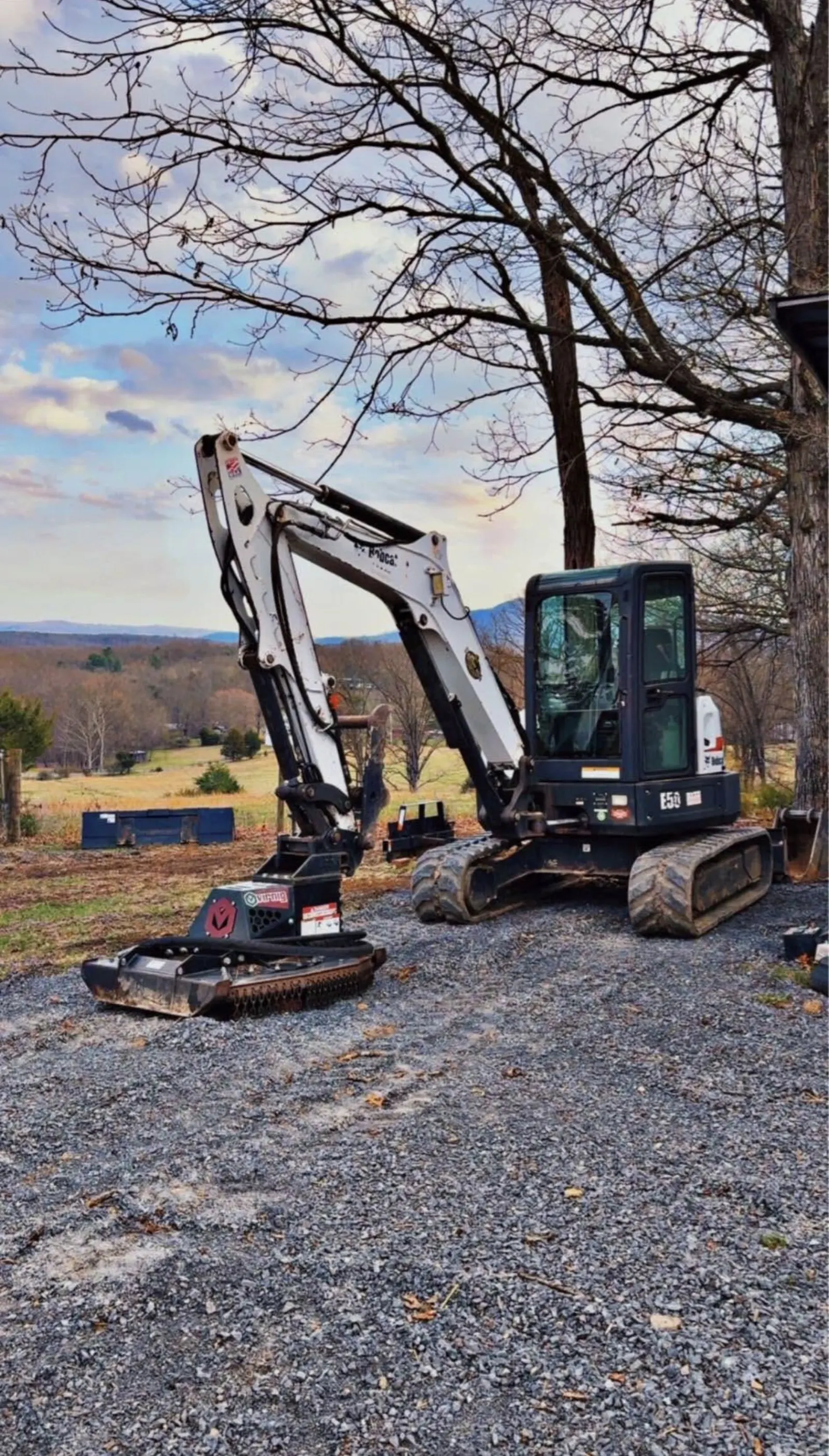 Mini excavator with a brush cutter head on a gravel pad, ridges in the distance
